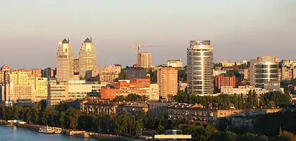 Vogelperspektive auf die Stadt bei Sonnenuntergang. Unten links ist ein Teil des Flusses zu sehen und mehrere Hochhäuser ragen aus dem Viertel hervor.