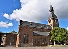 Seitliche Farbfotografie einer braunen Kirche mit einer Turmhaube an der rechten Seite, an der zwei Uhren zu sehen sind. Kleine Anbauten befinden sich um das Kirchenschiff, die lange Rundbogenfenster haben.