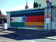 Seitliche Farbfotografie von der Berliner Mauer mit einem Bild, das aus drei Flaggen besteht. Links die palästinensische Flagge und rechts die deutsche Flagge und quer über beide befinden sich zwei blaue Linien und ein blauer Davidstern der israelischen Flagge. An den Seiten der Flagge stehen lange Texte und über dem Bild steht „März 90 Vaterland“. Links neben der Mauer ist ein Durchgang und im Hintergrund sind die Oberbaumbrücke und ein Haus zu sehen.