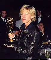 Porträtfoto von Ellen DeGeneres. Sie hält ihre linke Hand unter einen Emmy, den sie mit der rechten Hand festhält. Die goldene Statue sieht wie eine Frau mit Flügeln aus, die ein Atom-Modell in den Händen hält. DeGeneres trägt ein schwarzes Gewand mit weißen Blütenaufdrücken und ihre blonden Haare als etwas längere Bobfrisur mit Pony und nach hinten gekämmten Haaren. Hinter ihr sind ein an einem Tisch sitzender Mann und mehrere stehende Männer in Anzügen zu sehen.