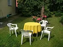 In einem privaten Garten auf grüner Wiese steht eine sitzgarnitur aus vier weißen Plastikstühlen und einem runden Tisch mit gelber Decke