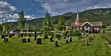 Foto einer dunkelbraunen Holzkirche mit rötlicherem Kirchturm. Hinter der Kirche befindet sich eine bergigere Landschaft.