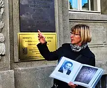 Gerlinde Kämmerer trägt schwarze Kleidung, hat blondes mittellanges Haar und hält ein Buch mit Fotos in der Hand. Sie steht vor einem grauen steinernen Gebäude, an dem eine goldene Gedenktafel angebracht ist.