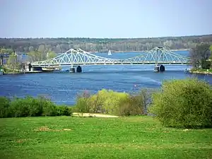 Farbfotografie von einer Brücke in einer Landschaft aus See und Wiesen mit Wald im Hintergrund. Die hellgrüne Stahlbrücke hat zwei Stützpfeiler.