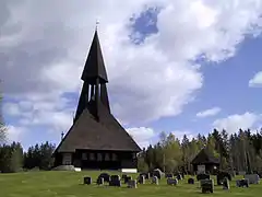 Foto einer dunkelbraunen Holzkirche mit einem Holzturm. Um die Kirche herum befindet sich ein Friedhof