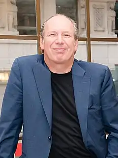 Ein Brustbild: Hans Zimmer in schwarzem Shirt und blauem Sakko. Er schaut zufrieden lächelnd in die Kamera, den Mund geschlossen. Er hat eine Stirnglatze.