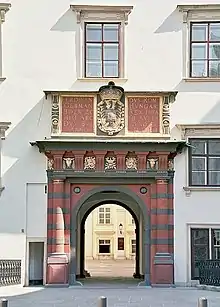 Frontale Farbfotografie von einem Durchgang in grün-braunen Querstreifen mit goldenen Ornamenten, der in die weisse Fassade mit Fenstern eingebaut ist. Über dem Durchgang befinden sich ein Wappen und lateinische Wörter.