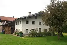 Farbfotografie von der Rückseite eines hellen zweigeschossigen Hauses mit Garten und Holzstapeln.