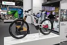 Ein weißes E-MTB steht auf einem Podest. Im Hintergrund ist eine Produktmesse zu sehen.