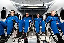 Vier Astronauten in blauen Overalls befinden sich in ihren Sitzen in einem Cockpit, und blicken in die Kamera.