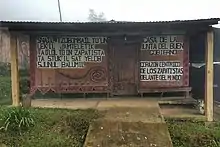 Holzbungalow mit rot-oranger Wandbemalung, Wellblech-Vordach und schwarzer Beschriftung auf weißem Grund sowie zapatistischen Ornamenten