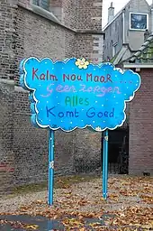 Ein blaues Schild, das die Form einer Wolke hat trägt in bunter Schrift die Worte: Kalm Nou Maal Geen Zorgen Alles Komt Goed. Im Hintergrund ist die Rückwand einiger Häuser zu sehen.