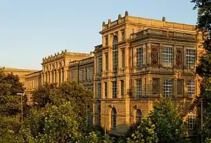 Seitliche Farbfotografie eines dreigeschossigen Gebäudes mit zwei Mittelteilen, die leicht vorgebaut und höher sind. Die hohen Fenster haben mehrere Sprossen und einige Rundbogenfenster befinden sich unter ihnen. Auf den Dächern stehen Steingeländer und Steinpfähle. An der rechten Seitenfassade sind zwischen den Fenstern hochkantige Reliefs. Das Gebäude wird von Bäumen umringt.