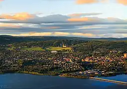 Foto einer Stadt, die an einem See liegt. Über den See führt eine Brücke. Hinter der Stadt erheben sich Hügel, an einer Erhebung befindet sich eine Skisprunganlage.