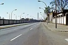 Seitliche Farbfotografie von einer Straße mit einer langen Mauer auf der linken Seite und einer Fabrikfassade auf der rechten Seite. Im Hintergrund fährt ein Auto und hinter der linken Mauer sind Häuser und ein Fabrikschornstein.