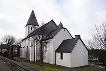 Von links nach rechts: Kirchturm, Hauptschiff mit Stützpfeilern, Chor (niedriger) und Sakristei (nochmals niedriger) der katholischen Pfarrkirche St. Margaretha Madfeld; alle Wände weiß gestrichen, schieferbedeckte Dächer. Fensterlose Rückansicht der Kirche mit Kirchhof, darauf laubfreie Bäume.