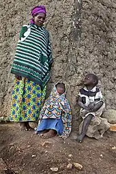 Eine ältere Frau mit Kleid und Kopftuch bekleidet steht vor dem Hauseck eines Lehmhauses und blickt lächelnd auf die zwei vor ihr sitzenden Kinder hinunter. Alle drei sind barfuß. Das rechte Kind sitzt auf einem Stein, die Hände locker auf dem überschlagenen Bein liegend und blickt zur Frau hoch. Das andere Kind hockt zwischen beiden auf dem Erdboden und hat sich in ein Tuch eingewickelt zwischen dem es nur mit einem Auge herausblickt.