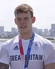 Farbfoto mit Richards unter freiem Himmel im weißen T-Shirt mit der Aufschrift Great Britain. Um den Hals trägt er ein rotblaues Band mit der Aufschrift Tokyo 2020, an dem seine Goldmedaille befestigt ist, die aber in diesem Bildausschnitt nicht zu sehen ist.