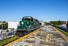 Dunkelgrüne Diesellok Nummer 300 mit weißen Beschriftungen NEW YORK AND ATLANTIC RAILWAY. Verfallener Mittelbahnsteig erhöht auf Viadukt/Bahndamm, funktionsloses Dach im Hintergrund, links und rechts leicht grün bewachsene Gleise und jenseits Gebäude der Stadt