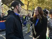 Aufnahme in einem herbstlichen Stadtpark. Zwei Personen groß im Bild: Laura Kavanagh, rechts, im Gespräch mit Jamie Torres-Springer, Abteilungsleiter der Metropolitan Transportation Authority, links. Daneben und im Hintergrund einzelne Zuschauer und Pressevertreter.