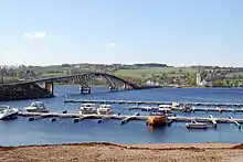 Foto einer Brücke über ein Gewässer. Auf der gegenüberliegenden Seite liegt eine Ortschaft mit einer Kirche. Vorne im Bild mehrere Boote an einer Marina.