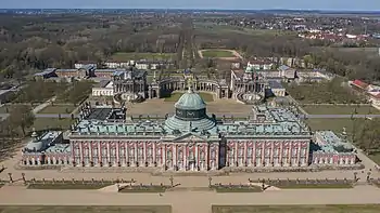 Frontale Farbfotografie in der Obersicht von einem Schloss mit vier Seitenflügeln und einer Dachkuppel. Auf den Dächern und vor dem Erdgeschoss sind Figuren aufgereiht. An den beiden äußersten Flügeln sind zwei kleine Dachkuppeln, die wie die großen Kuppelstatuen an der Spitze tragen. Im Hintergrund befindet sich ein halbrunder Säulendurchgang mit zwei Gebäuden, die Dachkuppeln und Säuleneingänge haben. Im Hintergrund sind weitere Gebäude und viel Wald mit Orten zu sehen.