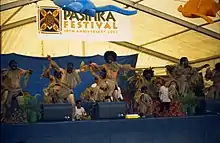 Tänzer aus Niue beim Pasifika Festival, 2002. Acht Männer in Gras-Rock-Bekleidung mit geschwärzten Gesichtern in ausdrucksvollen Posen.