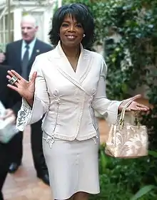 Porträtfoto von Oprah Winfrey. Sie trägt eine weiße Bluse, eine weiße Hose, einen silbernen Ohrstecker am linken Ohr und einen breiten, silbernen Ring am rechten Ring- und Mittelfinger. An ihrer linken Hand hängt eine hellbraun-weiße Handtasche mit Pflanzenabdrücken. Ihre schwarzen Haare trägt sie als bis zu den Ohren geschnittene Bobfrisur. Im Hintergrund sind verschwommene grüne Blätter und ein Mann im Anzug zu sehen.