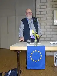 Tom Høyem in seiner Kluft als Wahlbeaobachter bei einem Vortrag des Mittwochsforums Rheinstetten 2024. Er steht hinter einem Tisch, auf dem Blumen stehen und an dem eine kleine EU-Fahne hängt.