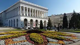 Farbfotografie mit Blick über ein rundes Blumenbeet zum klassizistisch erstellten Gebäude mit seinen weißen Säulen und Torbögen. Einige Tannen und ein weiteres Gebäude befinden sich auf der rechten Fotohälfte.