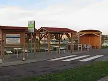 Gruppe von drei Holzdächern und mehreren Schildern ein einem schmalen, asphaltierten Weg. Unter zwei der Überdachungen sind einfache Bänke und ein Tisch aufgestellt, die hinterste hat eine Rückwand aus Holz, an der eine Bank angebracht ist. Zwischen den Überdachungen sind Anlehnbügel für Fahrräder aufgestellt. Im Hintergrund kann man einen Fluss und mehrere hell gestrichene kleinstädtische Häuser erkennen.