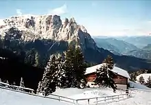 verschneite Schutzhütte, unten aus Stein, oben aus Holz erbaut; neben und direkt vor der Hütte Nadelbäume; im Vordergrund vor der Hütte die Fläche schneebedeckt; ein Weg mit einem teils im Schnee versunkenden Geländer abgetrennt; im Hintergrund Berggipfel aus blanken Fels; im fernen Hintergrund weniger hohe Berge, nicht schneebedeckt