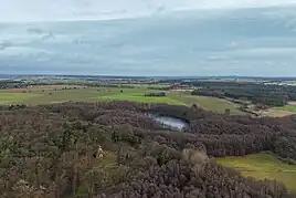 Ein Luftbild einer Naturlandschaft aus Wald, Wiesen und einem See, mit einer relativ kleinen, vierseitigen Natursteinpyramide mit hellen Eingangstor, die auf einer kleinen Waldlichtung inmitten eines Mischwald steht. Die Pyramide ist nicht höher als die Bäume.