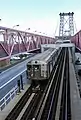 Blick vom Fußgängersteg auf die darunter verlaufende Brückenrampe der Williamsburg Bridge. Man blickt schräg von oben-vorne auf die Front und das Dach der ersten Wagen eines in gesicktem Edelstahl verschalten U-Bahn-Zugs. Die Brückenkonstruktion erscheint in einem bleichen Rotton, auf den Kraftfahrzeugfahrbahnen links und rechts ist wenig Verkehr sichtbar.