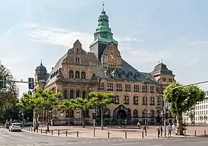 Frontale Farbfotografie eines dreigeschossigen Gebäudes mit einem Zwiebelturm in der Mitte und zwei Dachfassaden an beiden Ecken. Vor dem grünen Kupferturm sind eine Dachfassade mit einer Uhr und mehrere kleine Dachgiebeln zu sehen. Die linke Dachfassade hat drei Geschosse mit Rundbogenfenstern und die rechte Dachfassade besteht aus einem Geschoss mit rechteckigen Fenstern, die auch an der restlichen Fassade zu finden sind. Über dem Rundbogeneingang befindet sich ein Wappenrelief.