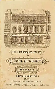 Revers CdV, Abbildung der Fensterfront des Ateliers (oberste Etage); davor Gitter mit Schriftzug „C. Seegert“; Atelier Carl Seegert, Berlin, Grosse Frankfurter Straße; Lithograf unbekannt; um 1880.