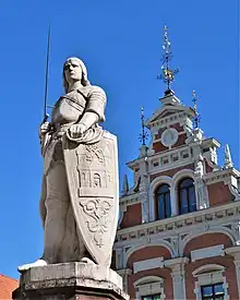 Seitliche Farbfotografie in der Untersicht einer weißen Statue mit Ritterrüstung und Wappenschild. Auf dem Schild befindet sich eine Burg mit Ornamenten. Im Hintergrund ist eine weiß-braune Staffelgiebel mit einem runden Kopfrelief und drei Eisenkreuzen.