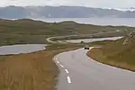 A road runs through a grassland tundra with the ocean in the background