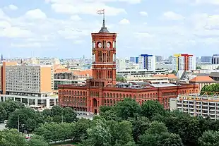 Frontale Farbfotografie vom Roten Rathaus mit seinen roten Backsteinen und Rundbogenfenstern. Das dreigeschossige lange Gebäude wird in der Mitte von einem hohen Glockenturm überragt, der an seinen Ecken mit Säulen verziert ist.