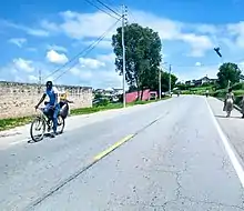 Eine einspurige asphaltierte Straße an der links Strommasten vorbeiführen. Auf der linken Straßenseite kommt ein Waren transportierender Radfahrer entgegen und auf einem Streifen neben der Fahrbahn sind rechts Fußgänger unterwegs. Im Hintergrund ist eine Ortschaft.