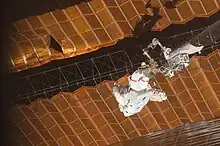 Two black and orange solar arrays, shown uneven and with a large tear visible. A crew member in a spacesuit, attached to the end of a robotic arm, holds a latticework between two solar sails.