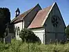 Eine steinerne Kirche mit einem steilen Ziegeldach. Im Bildvordergrund ist der Altarraum mit den runden Fenstern nach Osten zu sehen, im Hintergrund befindet sich auf dem höheren Kirchenschiff ein Glockenstuhl.