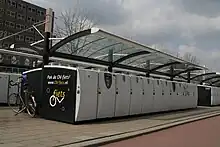 Farbfoto. Überdachte Gruppe Fahrradboxen. 2 Reihen mit je 12 Boxen, Rücken an Rücken. Bedienfeld an einigen Boxen. Seite beschriftet mit „Pak de OV-fiets! www.OV-fiets.nl“ und OV-fiets-Logo. Im Hintergrund eine zweite Gruppe Boxen.