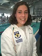 Tamsin Cook mit weißer Trainingsjacke in der Schwimmhalle. Die Trainingsjacke trägt die Aufschrift ihrer Universität.