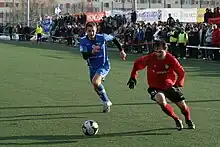 Frontale Farbfotografie von zwei Fußballspielern, die einem Ball in der Bildmitte hinterherjagen. Der linke Spieler trägt ein blaues Trikot und der rechte Spieler ein rotes Trikot und schwarze Torwarthandschuhe. Im Hintergrund steht ein kleines Publikum.