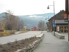 Foto einer Straße, die durch einen Ort mit mehreren Holzhäusern führt, dahinter eine Erhebung