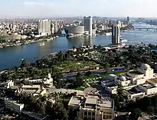 A sharp blue river divides the cityscape, which is primarily greenery and sparse low-rise buildings in the foreground, and dense with several modern high-rises in the background