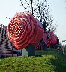 A large metallic sculpture of a red rose on a small grassy mound, with bare trees and other similar sculptures in the background