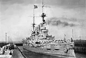 A large gray battleship sits in a lock, crew members in white uniforms crowd the ship's deck